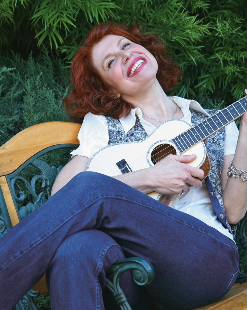 a photo of a red-haired smiling woman relaxing on a bench and strumming a ukulele