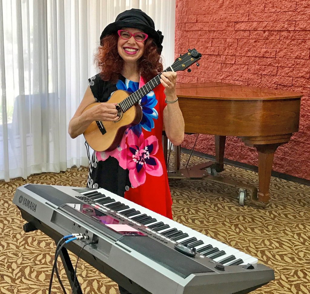 a woman plays a ukulele and stands in front of a keyboard