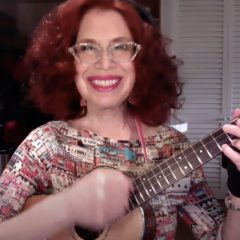 A red haired woman with glasses and a big smile plays a ukulele for the camera. Her hand strums the strings.
