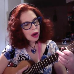 a woman with red hair and glasses plays the ukulele