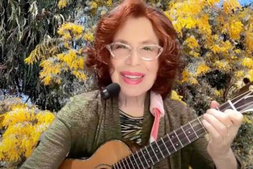 a woman with red hair and glasses plays a ukulele with yellow flowers in the background