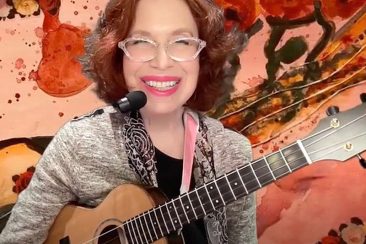a woman with red hair and glasses plays a ukulele with abstract patterns in the background