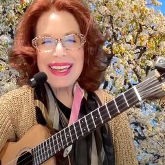 A woman with glasses, red hair, and colorful lipstick holds a ukulele in front of a background of flowering trees.