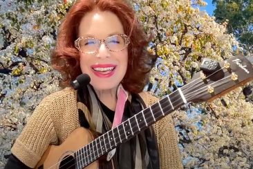 A woman with glasses, red hair, and colorful lipstick holds a ukulele in front of a background of flowering trees.