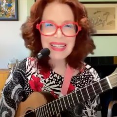 a woman with red glasses and a head microphone holds a ukulele